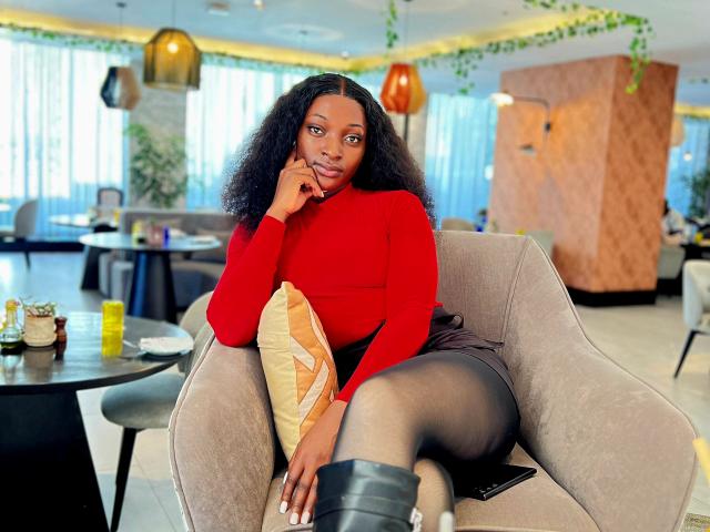 SexyShoku with curly black hair, red turtleneck, black skirt, and black boots, sitting in a beige chair, hand on chin, holding a yellow pillow, and a black phone.