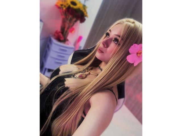 MiaValenttine with long blonde hair, pink flower in her hair, black lace dress, fair skin, looking up with a slight smile, wearing a necklace, in a bright room.