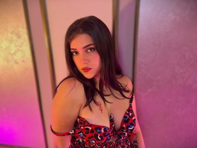 Katarina Novak, con i capelli scuri, indossa un abito a macchie di leopardo rosso e blu, che mette in mostra il seno, con rossetto rosa e una collana con un fiocco rosa, in una stanza illuminata di viola..