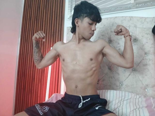Hombre musculoso sin camisa, con cabello negro, flexionando los bíceps, mostrando tatuajes en el brazo izquierdo, usando pantalones negros con cordón blanco, sentado en la cama, mirando hacia abajo, en una habitación con poca luz..