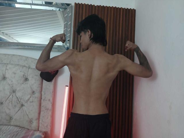 Shirtless SexlyMan with toned back and tattoos flexing arms, wearing black pants, standing in front of wooden panel and a mirror, showing off a muscular butt.