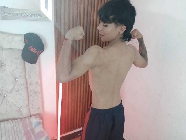 Um homem musculoso, sem camisa, com cabelo preto, exibindo seus braços, mostrando suas costas definidas e um braço tatuado, vestindo calças escuras, em pé em um quarto, usando um chapéu..