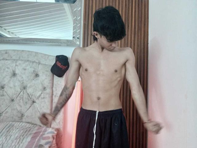 Hombre sin camisa, "SexlyMan", con cuerpo delgado y tonificado, pechos pequeños, cabello negro, pantalones cortos negros, tatuaje en el brazo derecho, de pie con los brazos extendidos, mirando hacia abajo, pared de madera detrás de él..