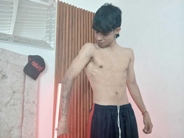 Hombre sin camisa, llamado "SexlyMan", con cabello negro, aretes en las orejas, tatuajes y pantalones negros, de pie con los brazos ligeramente flexionados, mirando hacia abajo, en una habitación con una luz vertical roja..
