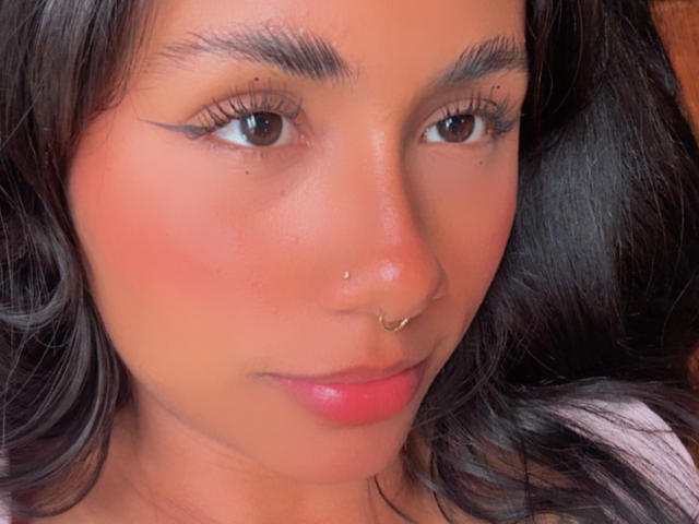AnaRome has brown eyes, a small nose ring, and a subtle smile. Her pink lips and slightly flushed cheeks are visible. She has dark, wavy hair and well-groomed eyebrows.