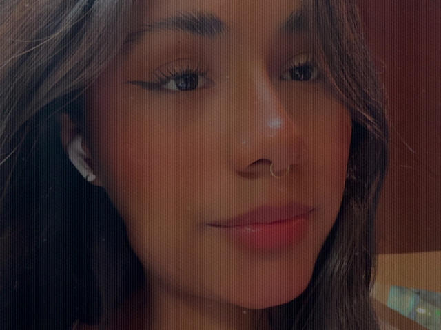 AnaRome's face with brown wavy hair, black eyeliner, nose ring, and white earbuds, smiling slightly with pink lips. Soft lighting, gentle focus.