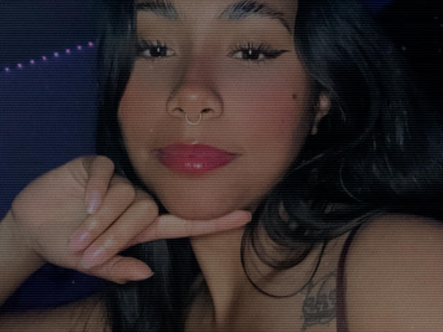 AnaRome's face with dark hair, nose ring, and pink lipstick, hand under chin, wearing a sleeveless top, showing a tattoo on her right shoulder, slightly blurred.