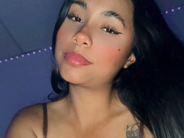 AnaRome with long black hair, nose ring, and thick eyeliner, pouty pink lips, small tattoo on right shoulder, wearing brown strap, soft purple lighting.