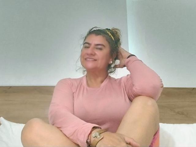 FuegoSexy, a curvy woman with brown hair in a messy bun, wearing a pink long-sleeve top, sits with knees up, smiling, hand in hair, on a bed.