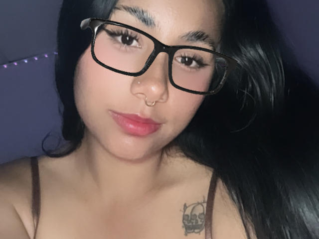 AnaRome with black glasses, nose ring, pink lipstick, and long black hair, showing a tattoo on her right shoulder, looking at the camera, with a slight pout.