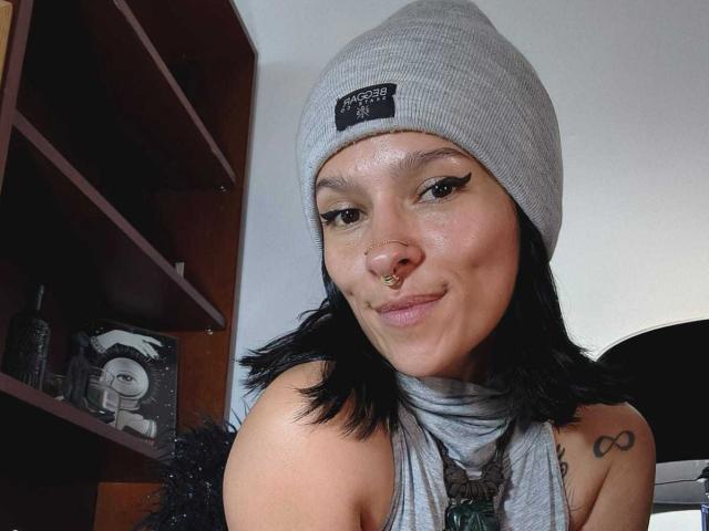 AmelieBlake with black hair, grey beanie, nose ring, and black eyeliner, smiling at camera, showing bare shoulders with small tattoo on right side, wearing grey scarf.