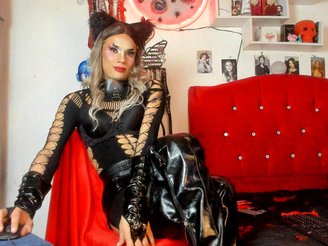 MistressHuggecock with cat ears, black lace-up bodysuit, red skirt, black leather boots, and caged collar, sitting on red tufted bed, looking at camera. Her tits are visible.