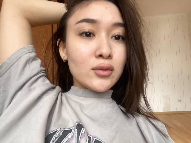 IzettaDingel has puffy lips, brown eyes, and straight brown hair, wearing a grey t-shirt with one arm raised, showing a nose ring and small earrings.