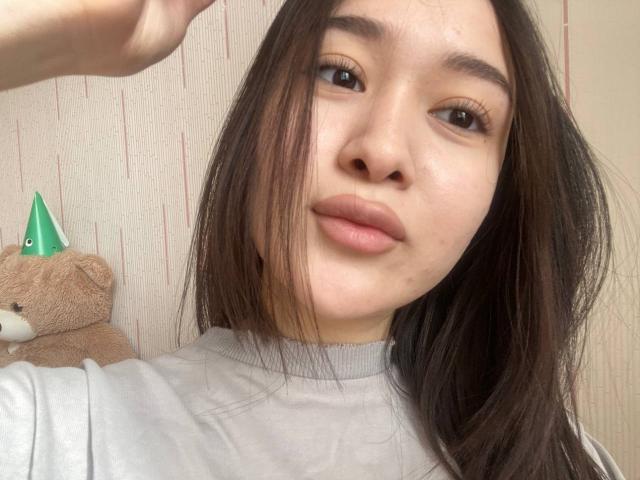 IzettaDingel with puffy brown eyes, straight brown hair, and light skin, wearing a grey shirt, has a cute face and a small pimple on her cheek. Teddy with green hat.