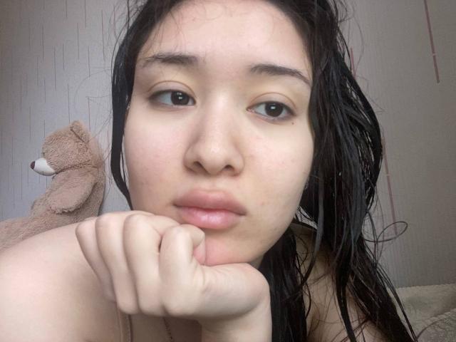 IzettaDingel with wet black hair, pouty lips, and hand under chin, brown eyes, light skin, small nose, looking at camera, soft teddy bear on left.