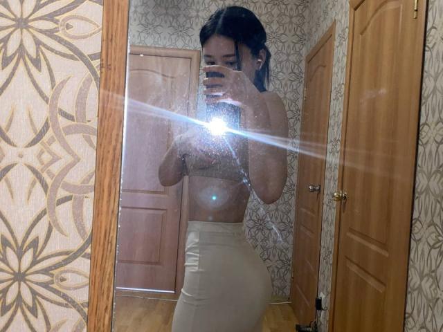 IzettaDingel in a mirror selfie, wearing a brown sports bra and tight beige leggings, showing her ass and small tits, holding a phone with a bright flash.