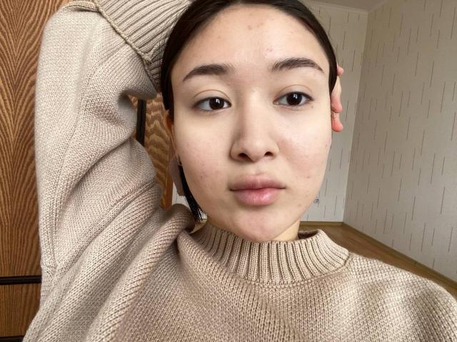 IzettaDingel's face, with light skin, dark eyebrows, and brown eyes, wearing a beige knitted sweater, has a hand in her hair, looking slightly bored. Her tits are small.