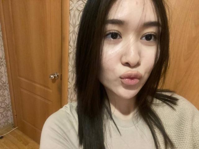 IzettaDingel with puffy, glossy lips, small nose, and straight black hair, wearing a beige top, has a cute, slightly annoyed expression, in a wooden room.