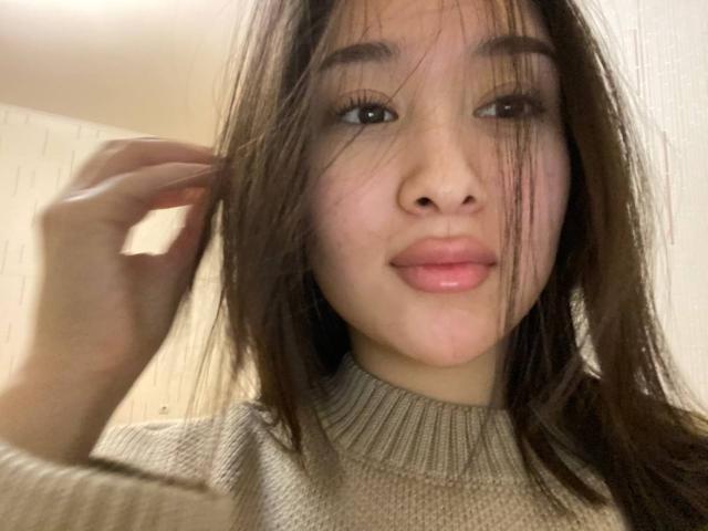 IzettaDingel with long brown hair, slightly messy, wearing a beige turtleneck, holding her hair with one hand, and a soft, natural smile, pinkish lips, and subtle makeup.