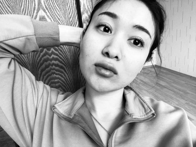 Black-and-white photo of IzettaDingel with pouty lips, dark eyebrows, and messy hair, wearing a zip-up jacket, hand behind head, wooden floor, and textured wall in background.
