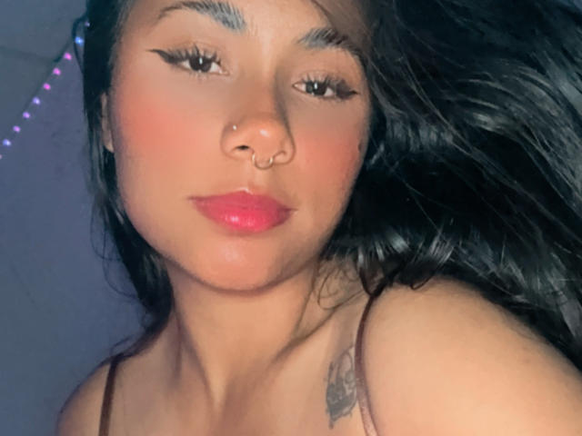 AnaRome with pink lips, nose ring, and black eyeliner, looking at the camera, showing her tattoo on the right shoulder, and dark, wavy hair, in a selfie.