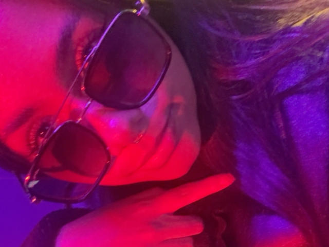AnaRome with pinkish-red neon lighting, wearing dark sunglasses, showing a middle finger with her right hand, has long brown hair, and a slight smile, looking directly at the camera.