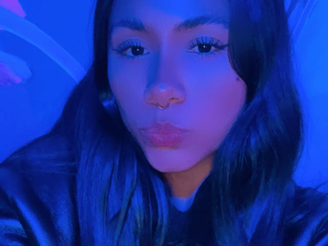 AnaRome's face in blue light, pouty lips, long eyelashes, dark hair, subtle nose, slightly tanned skin, wearing a black shirt, looking directly at the camera.