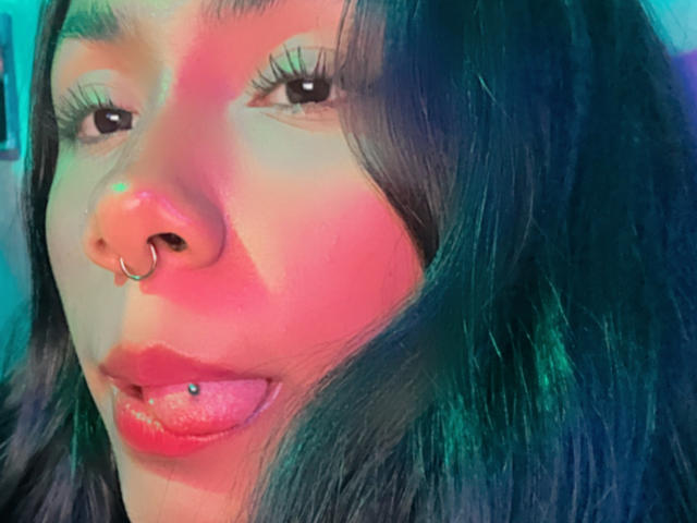 AnaRome has a nose ring, tongue piercing with a blue ball, and dark, wavy hair. Her face is lit with colorful, neon-like lighting.