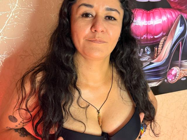 DanyUmil has curly black hair, tan skin, and wears a black bra with a pendant. She's looking at the camera with a subtle smile, showing small tits.