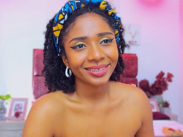 MeganWilliams with curly black hair, yellow-green eye makeup, nose ring, silver hoop earrings, and blue-yellow patterned headband, smiling at the camera, topless, showing her small, cute tits.