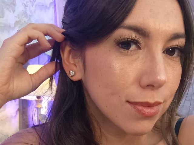 MiaReed's face, slightly smiling, with brown hair, holding her hair up, showing a small diamond stud earring, and wearing pink lipstick, showing her ass-kissing glossy lips.