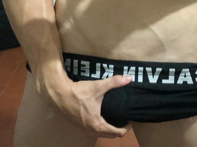 La main de SebasSexyMan serre ses sous-vêtements noirs, les tirant vers le bas pour révéler son pénis circoncis et flasque. Sa peau est pâle, et sa main est légèrement musclée..