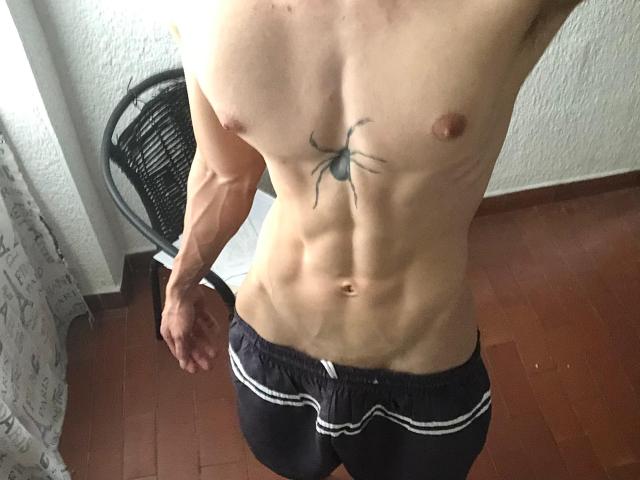 Le torse musclé et nu de SebasSexyMan, avec un tatouage de toile d'araignée noire entre de petits mamelons bruns, portant un short noir à rayures, ample, debout dans une pièce avec des planchers en bois..