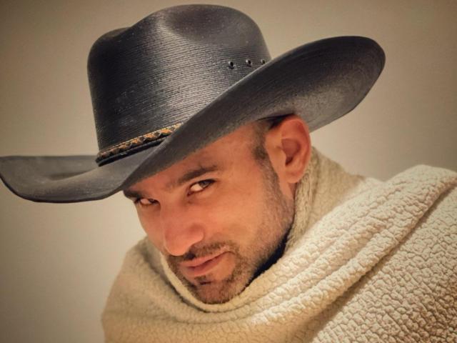 PrimeDominus, com um chapéu de cowboy texturizado em cinza escuro e um cachecol branco de tricô, olhando de lado com um leve sorriso, exibindo uma barba bem cuidada e olhos castanhos..