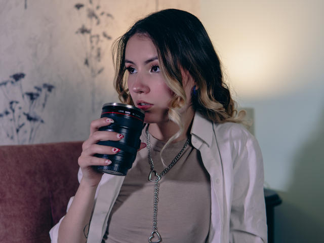 LilaLaurent with blonde and brown wavy hair, holding a black metal can, wearing a beige top, white shirt, and chain necklace, looking thoughtful with red nail polish.