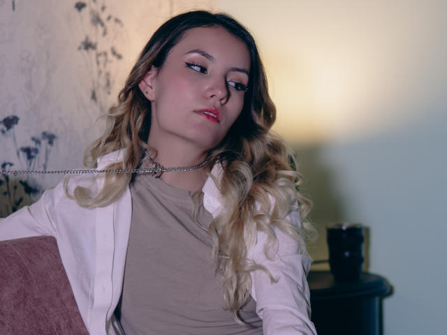 LilaLaurent, con capelli biondi ondulati, camicia bianca e top beige, seduta, con un'espressione leggermente annoiata, una catena al collo, rossetto rosso, illuminazione soffusa e una lampada sullo sfondo..