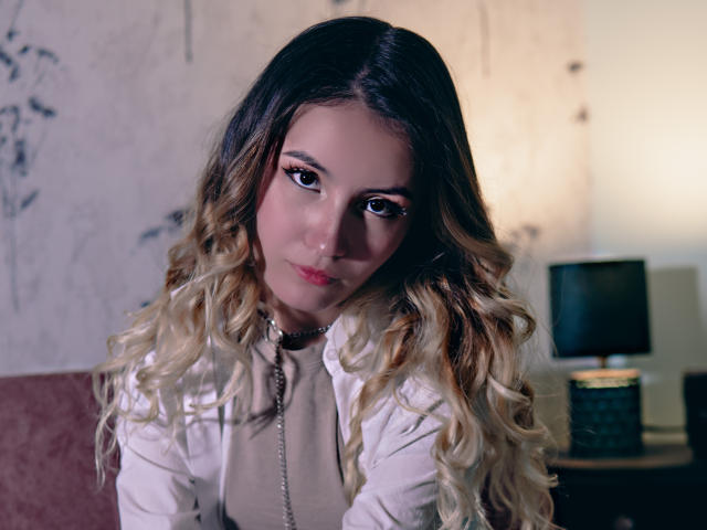 LilaLaurent has long, wavy blonde and brown hair, wearing a white shirt, looking at the camera with serious eyes, and a black choker. Her tits are small and cute.