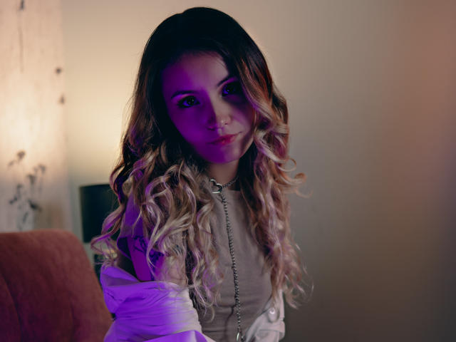 LilaLaurent with long, wavy, blonde-tipped hair, purple-lit face, and tattooed arm, wearing a white top, and a chain necklace, sitting with a subtle, confident smile.