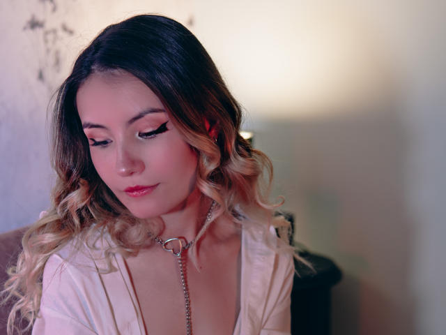 LilaLaurent with wavy blonde hair, wearing a white shirt and silver chain collar, has pink blush and black eyeliner, looking down with a slight smile, in a softly lit room.