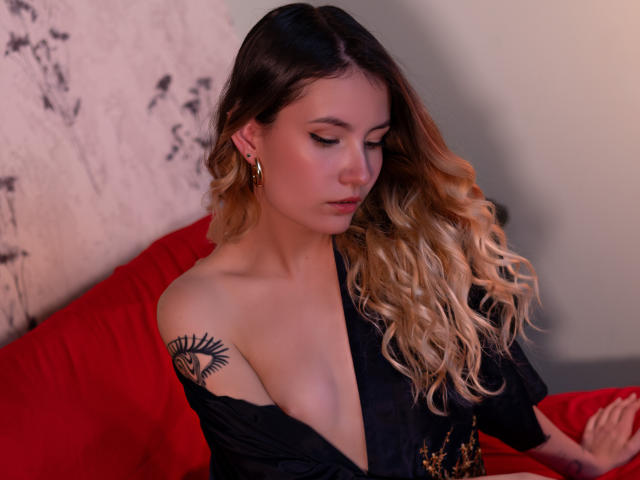 LilaLaurent with blonde, wavy hair, black off-shoulder top, gold earrings, and eye tattoo on her shoulder, sitting on red couch, looking down, soft pink and warm light.