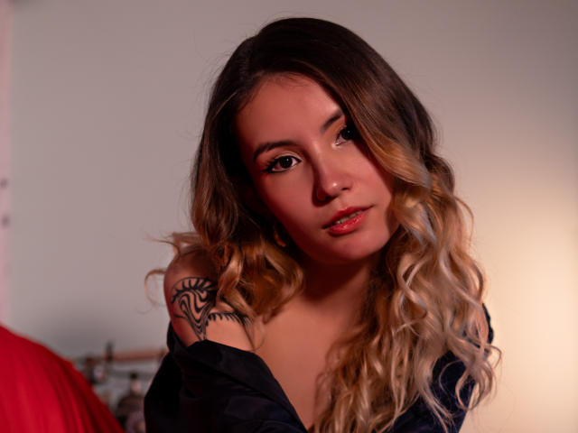 LilaLaurent with wavy blonde and brown hair, dark tattoo on right shoulder, wearing black off-shoulder top, soft light, looking down, red bed in background, subtle makeup, cute.
