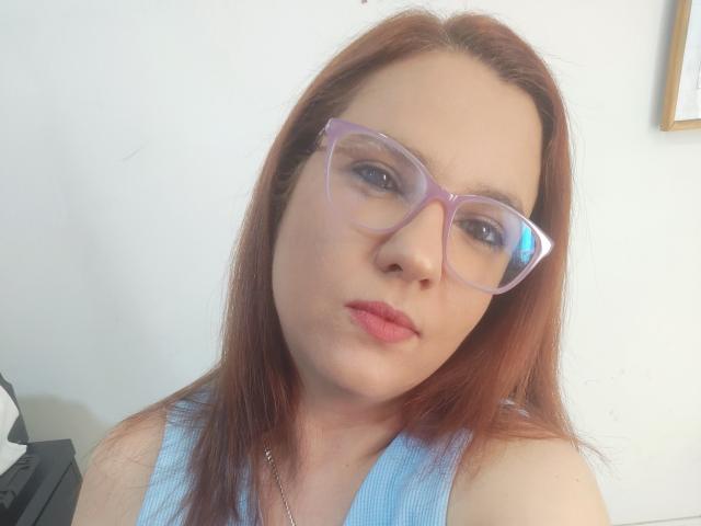AmeliaWilson with red hair, purple glasses, and pink lips, wearing a blue sleeveless top, has a serious expression, showing her face and upper chest.