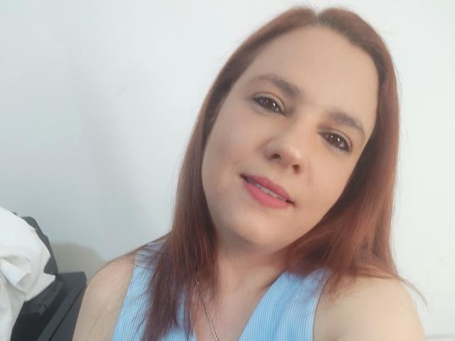 AmeliaWilson has red hair, light skin, brown eyes, and pink lipstick. She's wearing a blue sleeveless top with a silver necklace. Her tits are small and she's smiling.