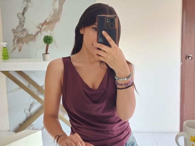 MiaReed with long dark hair, wearing a purple tank top, taking a mirror selfie with her phone, showing her tits, wearing bracelets, in a bright room.