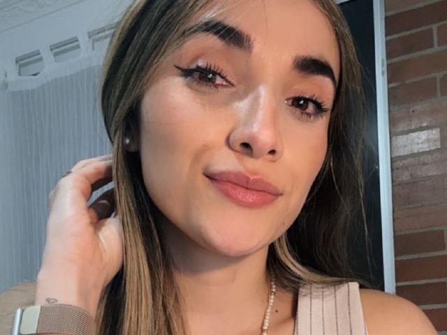 AdharaCooperr's face with light brown hair, thick eyebrows, long eyelashes, pink lips, and a slight smile, wearing a white top, and a pearl necklace, with her hand near her ear.