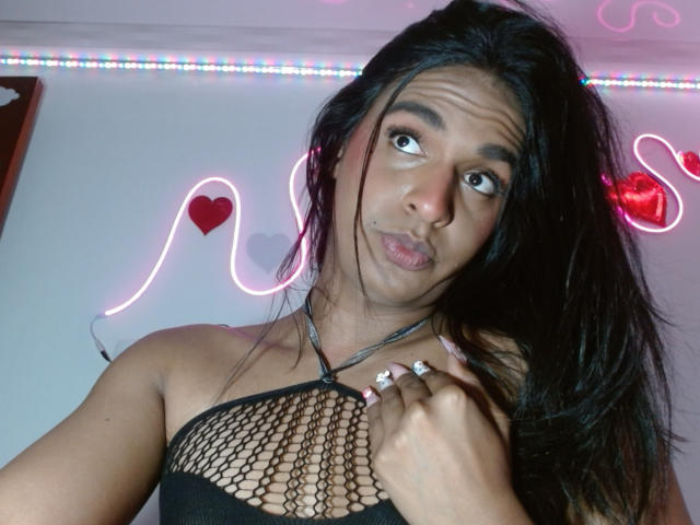 IdiiaLatina with dark long hair, pouty face, black fishnet top showing tits, and a ring on her finger, against neon heart and snake wall decor.