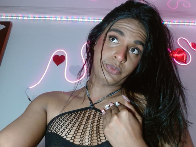 IdiiaLatina with long black hair, wearing a black mesh top, holding her chest, pouty face, pink neon heart and snake lights in background, colorful LED strip above.