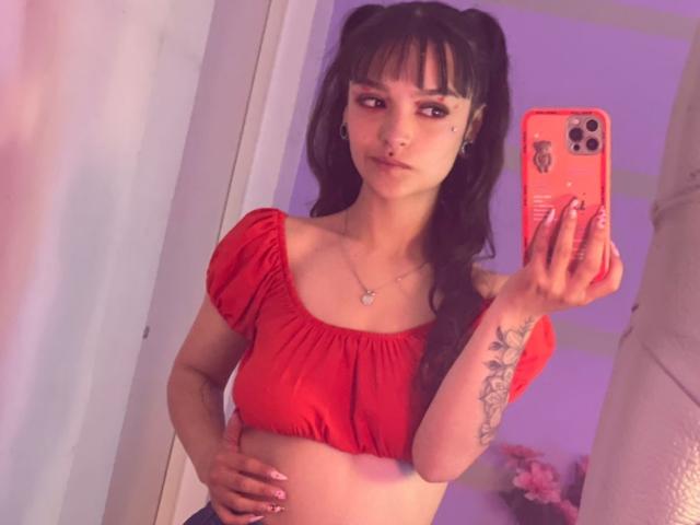 MarietteJones with pigtails, red crop top, showing her tits, holding a pink phone, tattoo on right arm, purple wall, hand on hip, looking in mirror.