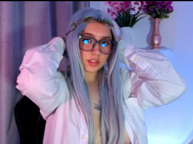 Susan Monroy, con cabello largo y plateado, usando gafas, camisa blanca, manos en el cabello, ojos azules, diadema con flores, jarrón rosa con flores en el fondo, iluminación suave en tonos azul y rosa..