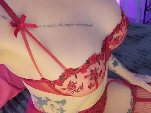ElixirShanna's red lace bra with floral embroidery, showcasing her tits and tattoos, including a cat on her hip. She's wearing matching red lingerie, sitting on a bed.